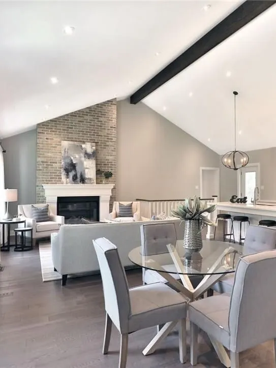 An open-concept dining and living area under a vaulted ceiling with a dark beam and a brick fireplace