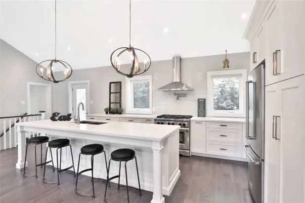 A finished white kitchen with a long island, four black stools, two round pendant lights and stainless appliances.