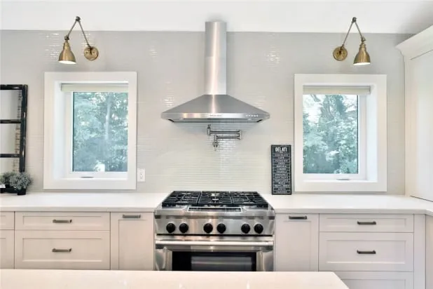 A kitchen wall with a gas range and chimney hood centred between two windows under gold wall lamps.