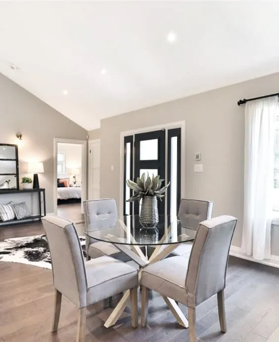 A round glass table and four grey upholstered chairs in the dining area, near the black front door.