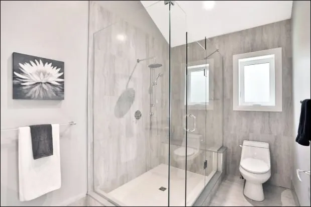 A bathroom with a glass shower, grey tiled walls, a rain shower head and a white toilet.