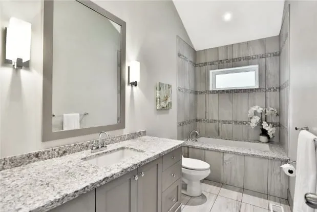 A finished bathroom with a grey vanity, speckled countertop, large mirror and a tiled tub with mosaic bands.