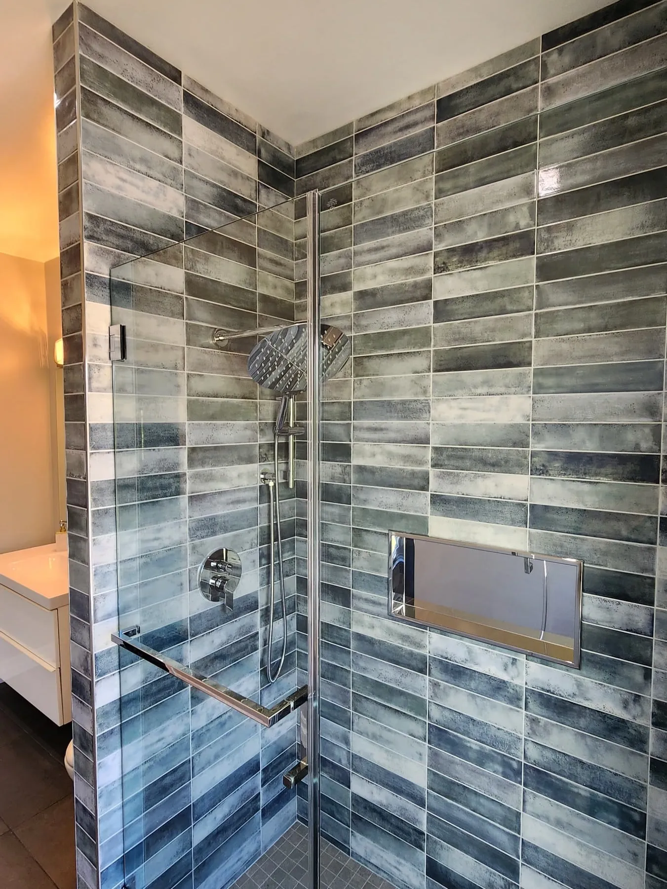 A glass-enclosed shower with long grey and blue tiles, a rain shower head and a built-in niche