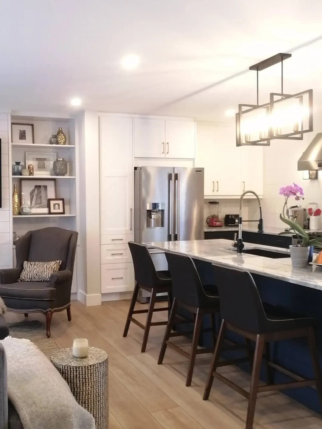 An open kitchen with a long island and black stools beside a living area with built-in shelves